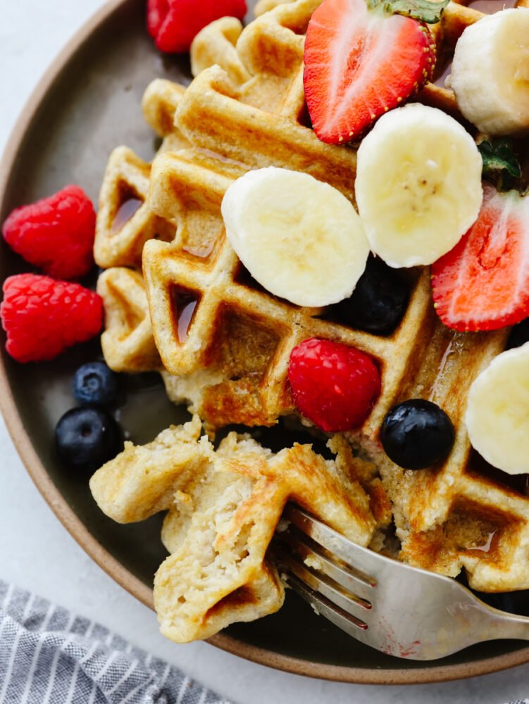 Zoomed in shot of a waffler on a plate. It's topped with fruit and being cut into with a fork. 