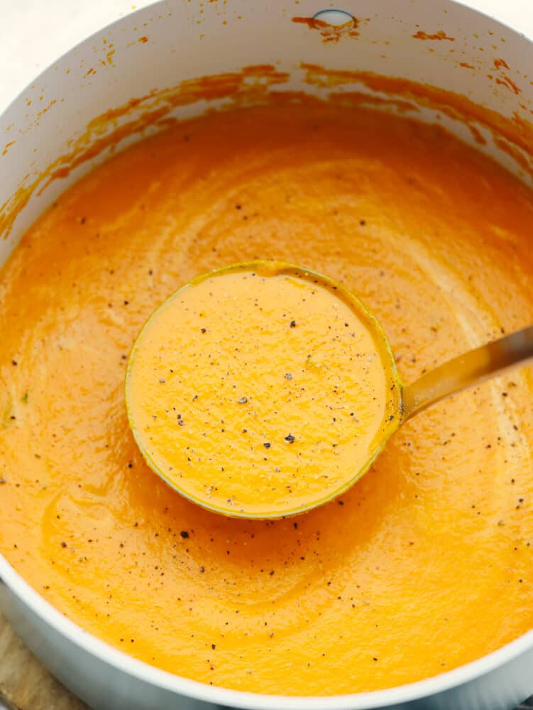 A pot of carrot soup sprinkled with pepper and a ladle scooping some out. 