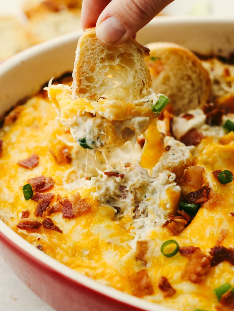 A close up of crack chicken dip with a small piece of bread being dipped into it. 