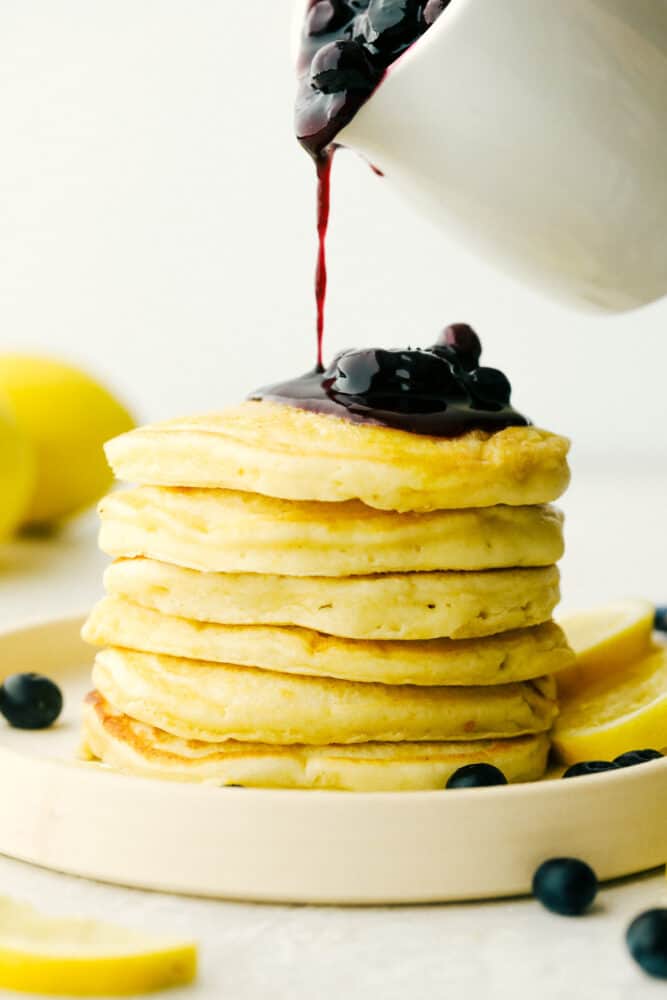Pouring blueberry sauce over a stack of lemon pancakes.