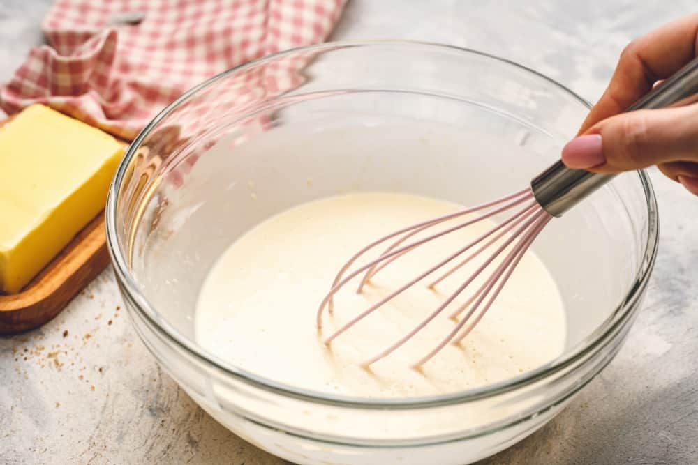 Cinnamon roll pancake mix being stirred together with a whisk in a glass bowl with a side of butter.
