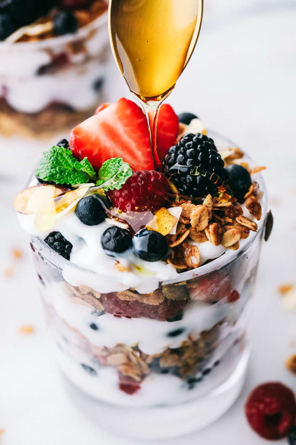 yogurt parfait in a glass cup with honey being drizzled on by a spoon. 