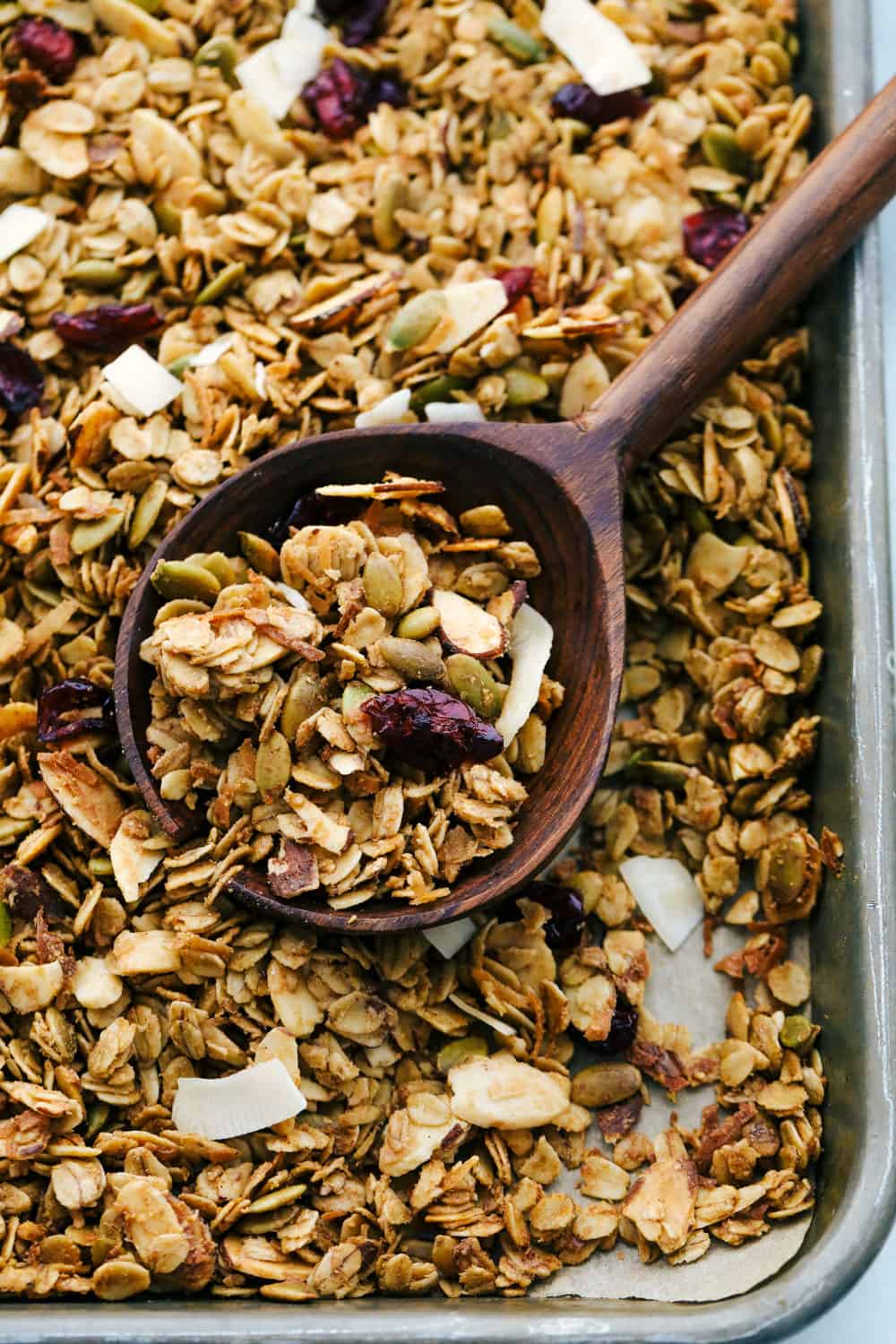 Homemade baked granola on a sheet pan with a large wooden spoon scooping it up. 