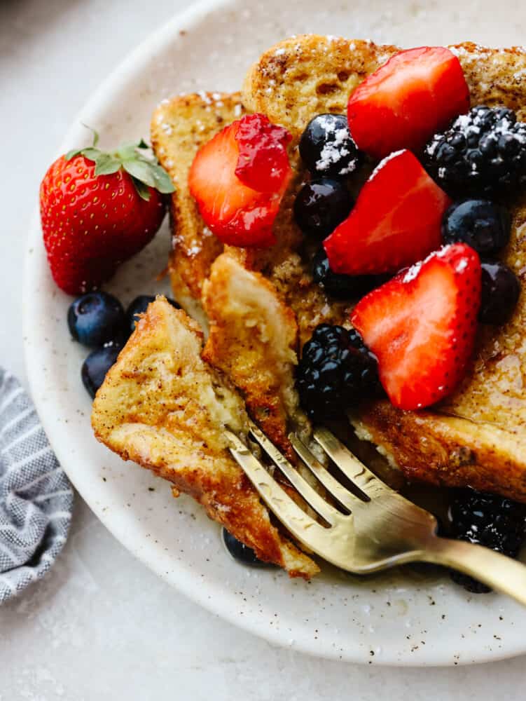 A piece of french toast on a plate with berries and syrup, being cut into with a gold fork. 