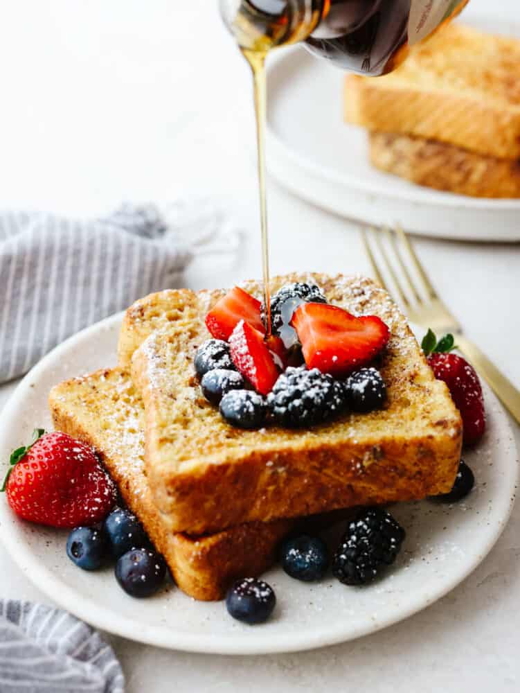 2 pieces of french toast on a plate with berries and powdered sugar with syrup being poured on top. 