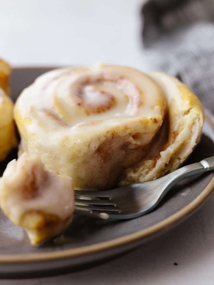 A cinnamon roll on a plate with a silver fork cutting into it. 