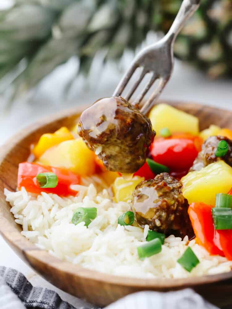 A wooden bowl filled with rice, meatballs and vegetables. One meatball is being poked with a fork. 