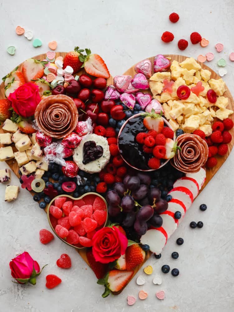Meats, cheese, fruit and candy on a heart shaped wood cutting board.