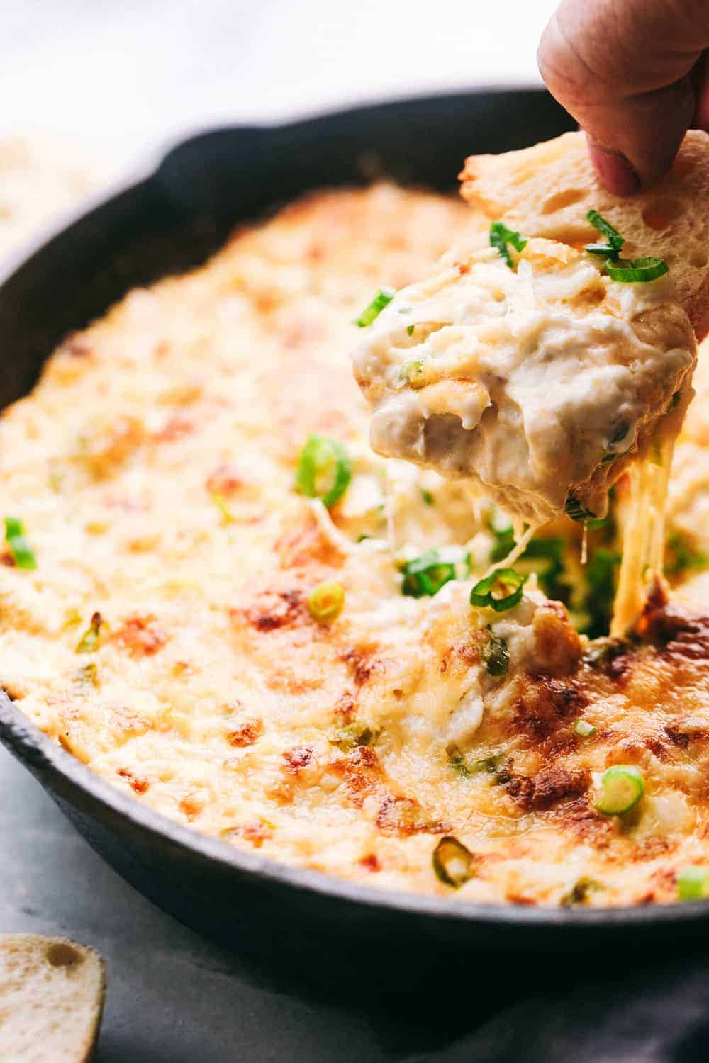 Hot Crab Dip in a metal pan. There is piece of bread dipping into the pan. 