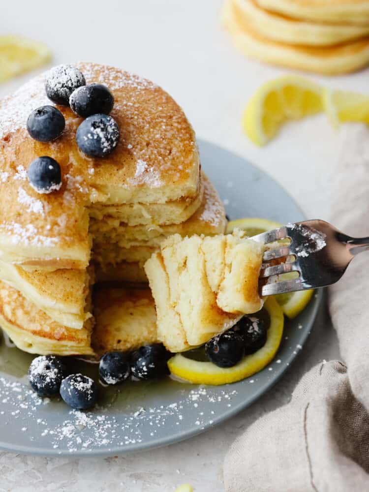 A stack of pancakes with blueberries and lemons and powdered sugar. They are being cut into with a silver fork. 